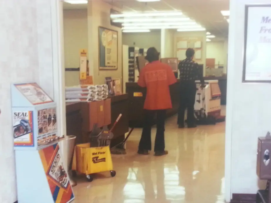 Two AMS crew members in red AMS jackets cleaning a store floor beside a wet floor sign
